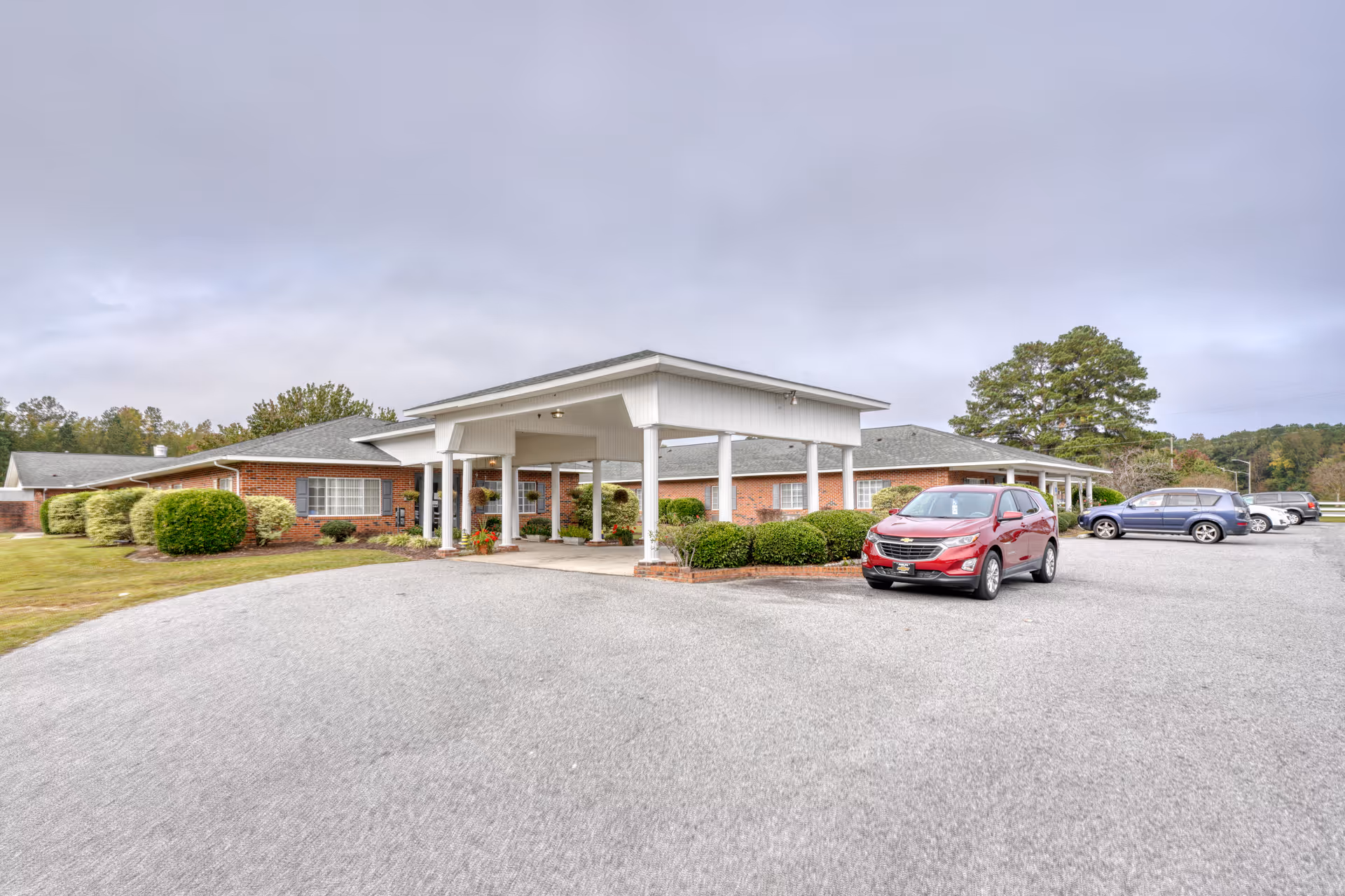 Exterior view of a single-story brick building with a covered entrance supported by white columns. Several cars are parked in the gravel parking lot in front of the building. The surrounding area has green bushes, grass, and trees under a cloudy sky.