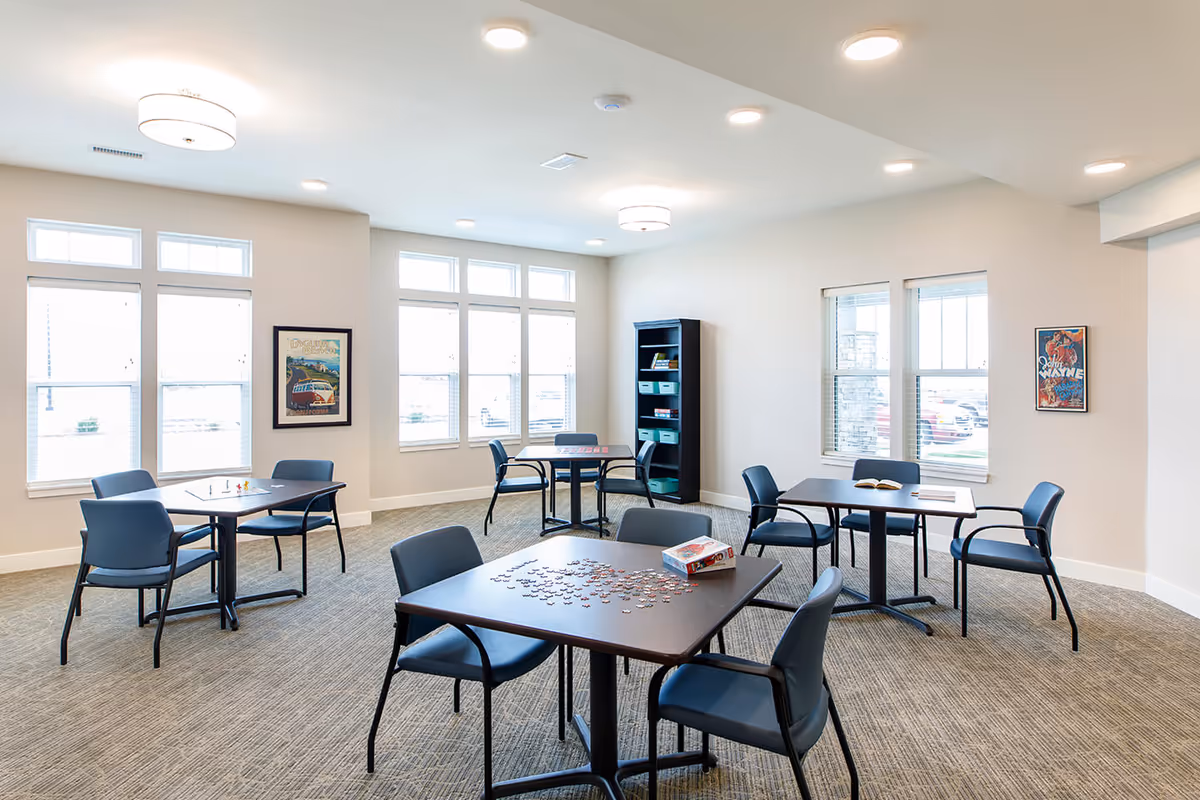 Well-lit common room with multiple tables and chairs, a jigsaw puzzle on one table, large windows, and a bookshelf.