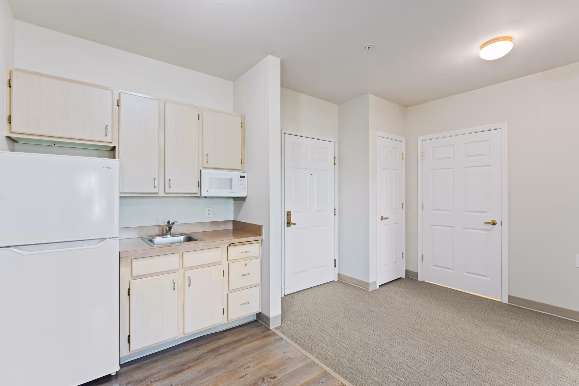 A small kitchen area with light wood cabinets, a white refrigerator, a microwave mounted above the countertop, and a stainless steel sink. The kitchen is adjacent to a carpeted area with three white doors, one of which appears to be an entrance door. The walls are painted white and the floor near the kitchen is wood laminate.