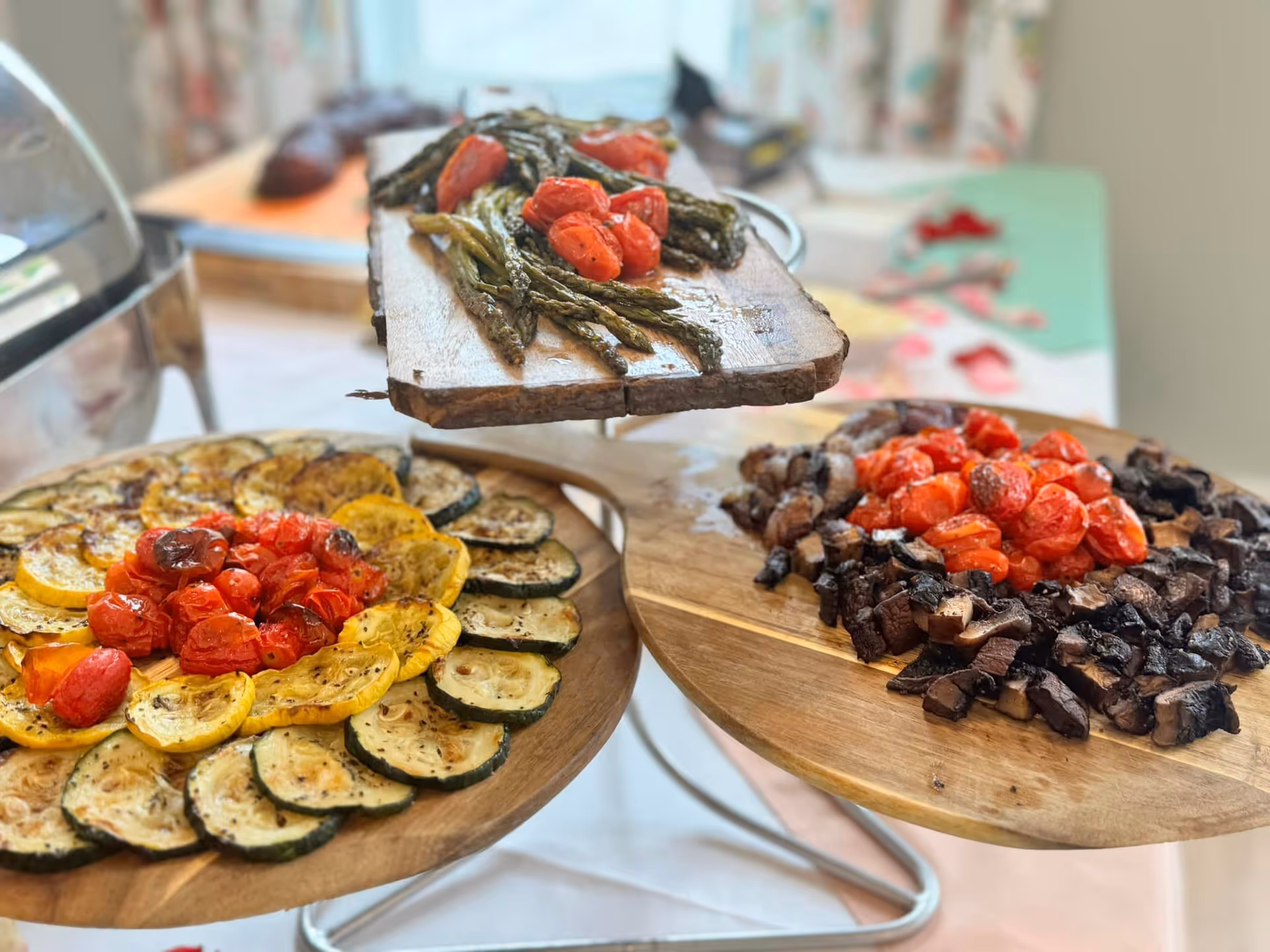 Three wooden serving platters with roasted vegetables including asparagus and cherry tomatoes on the top platter, sliced zucchini and yellow squash with cherry tomatoes on the left platter, and chopped mushrooms with cherry tomatoes on the right platter, displayed on a table with a blurred background.