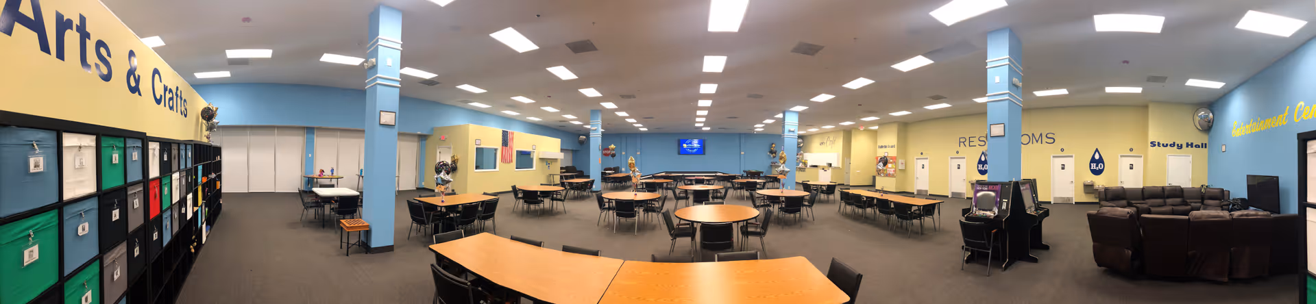 A large, well-lit multipurpose room with tables and chairs arranged for group activities. On the left side, there is a shelving unit labeled 'Arts & Crafts' with colorful storage bins. The room has blue pillars and yellow walls with various signs including 'Restrooms,' 'Study Hall,' and 'Entertainment Center.' There are several arcade machines and a seating area with brown recliners on the right side.