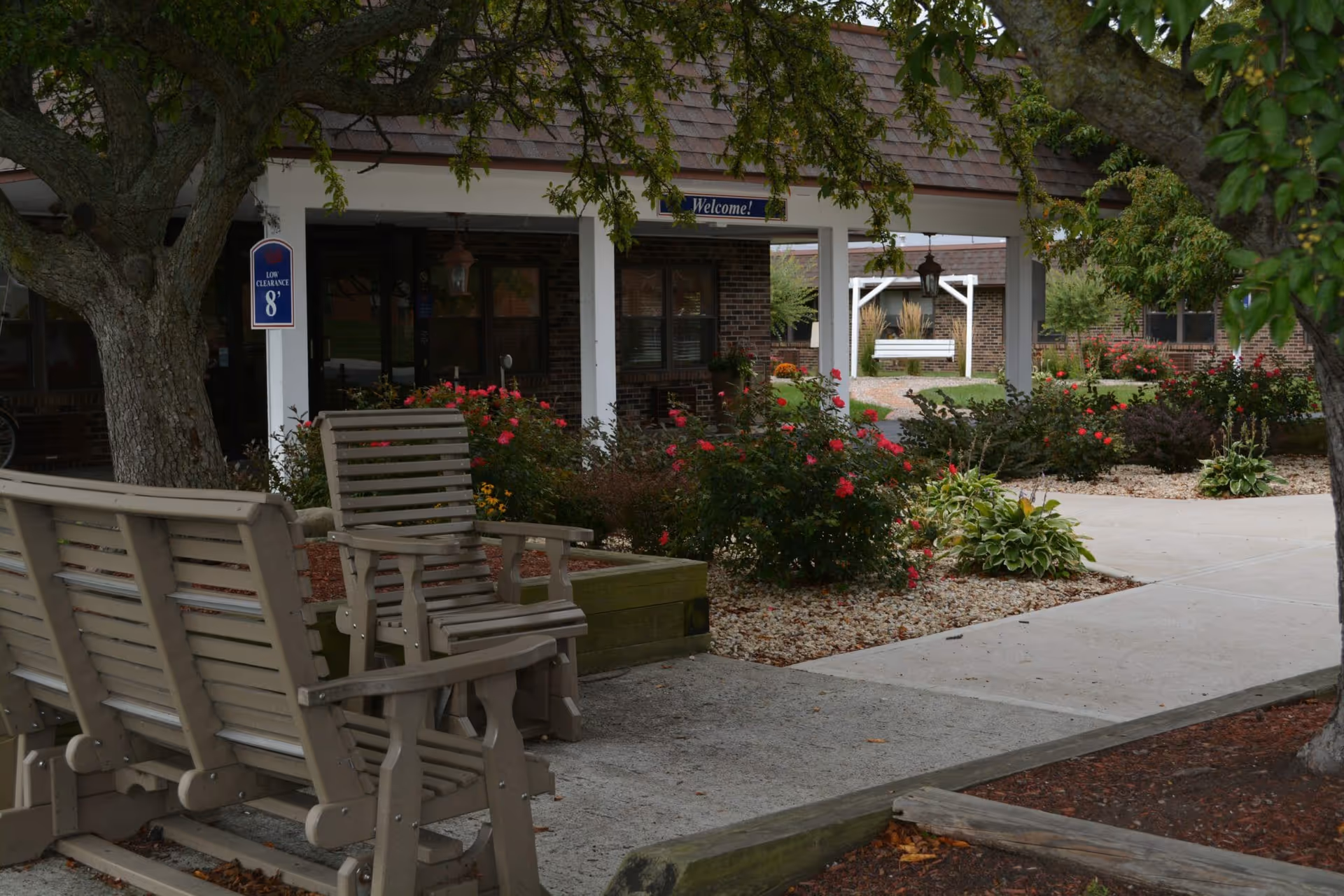 Courtyard with wooden benches and chairs, flower beds, and a covered entrance to a care facility.