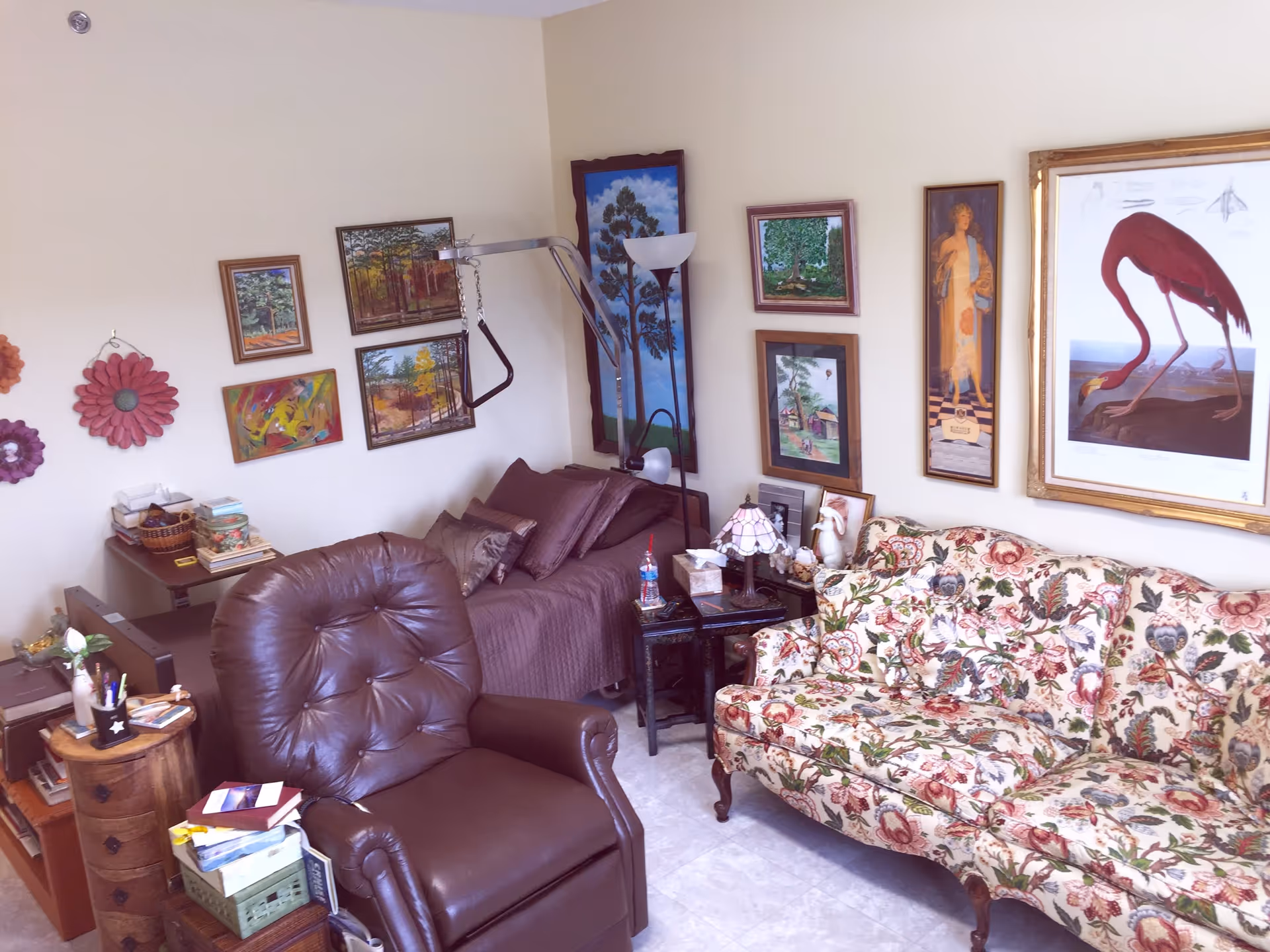 A cozy living room area with a brown leather recliner, a floral patterned sofa, and a bed with brown bedding. The walls are decorated with various framed paintings and artwork. There is a small side table with a lamp and decorative items next to the bed.