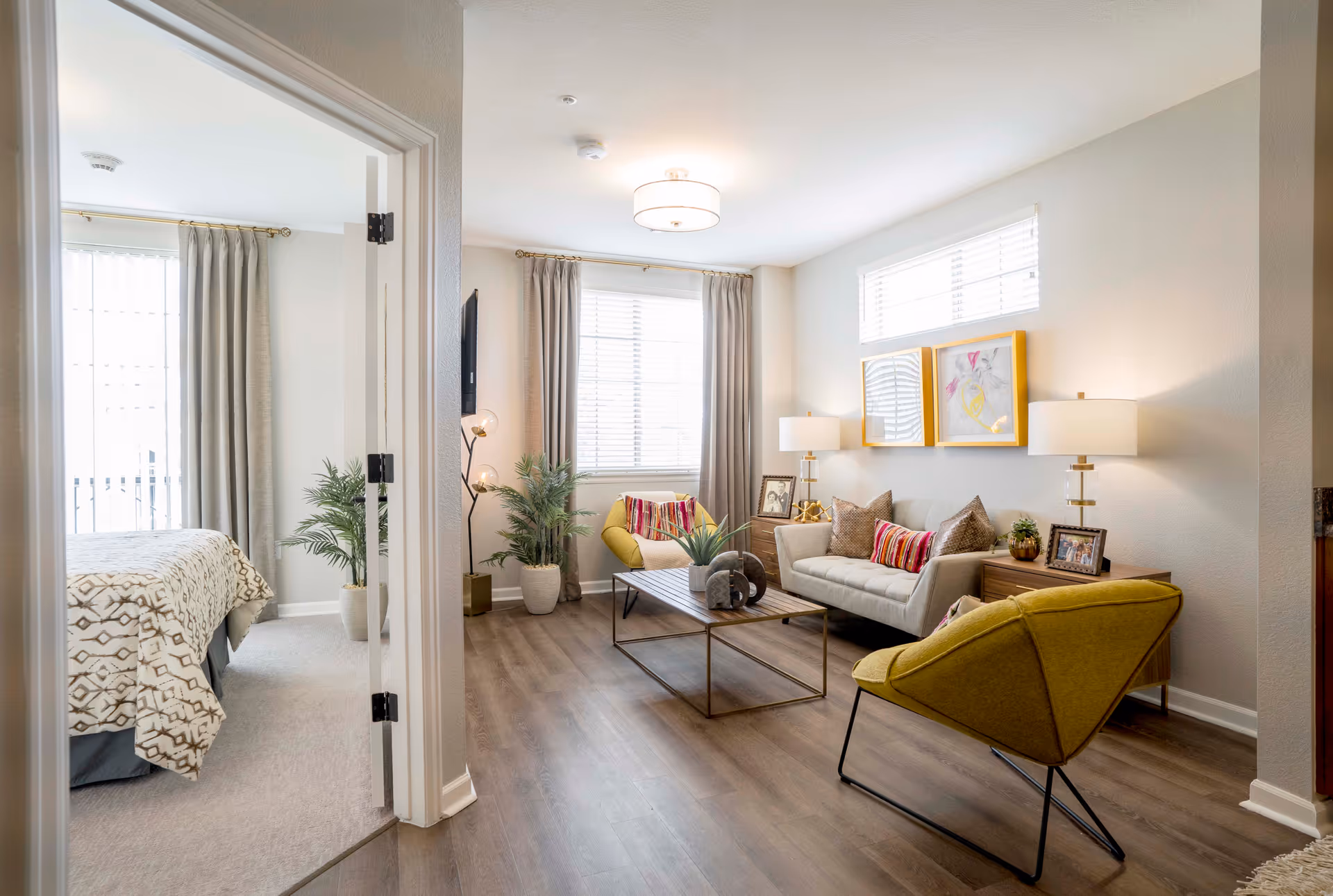 A bright and modern living room with a beige sofa adorned with colorful pillows, two yellow armchairs, a wooden coffee table with decorative items, and two side tables with lamps and framed photos. The room has large windows with curtains, light wood flooring, and neutral-colored walls. To the left, an open doorway reveals a bedroom with a bed covered in a patterned blanket and a large window with curtains.