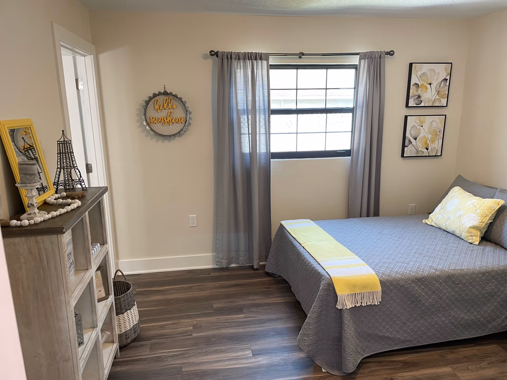 A bedroom with a bed covered in a gray quilt and a yellow and white throw blanket. There is a yellow pillow on the bed. Two framed floral artworks hang on the wall above the bed. A window with gray curtains is centered on the wall. To the left, there is a wooden shelf with decorative items including a small Eiffel Tower model, a yellow picture frame, and a beaded garland. A round wall decoration with the words 'hello sunshine' is hung on the wall near the shelf. The floor is wood with a dark finish.