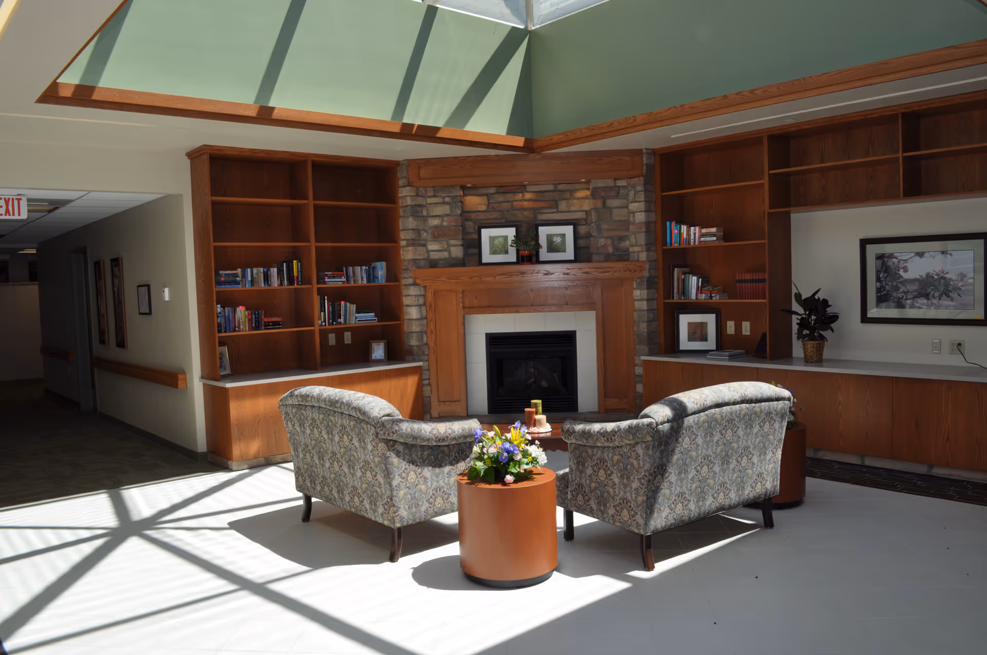 A cozy sitting area in a senior living facility with two patterned armchairs facing a stone fireplace with a wooden mantel. The room has built-in wooden bookshelves filled with books and framed pictures. A small round table with a flower arrangement is placed between the chairs. Sunlight streams in through a skylight, casting shadows on the tiled floor.