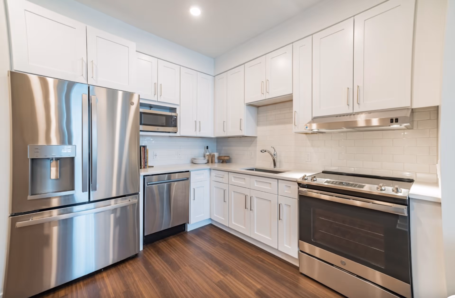 Modern kitchen with white cabinetry, stainless steel refrigerator, dishwasher, microwave, oven, and a sink. The kitchen has a white subway tile backsplash and wooden flooring.