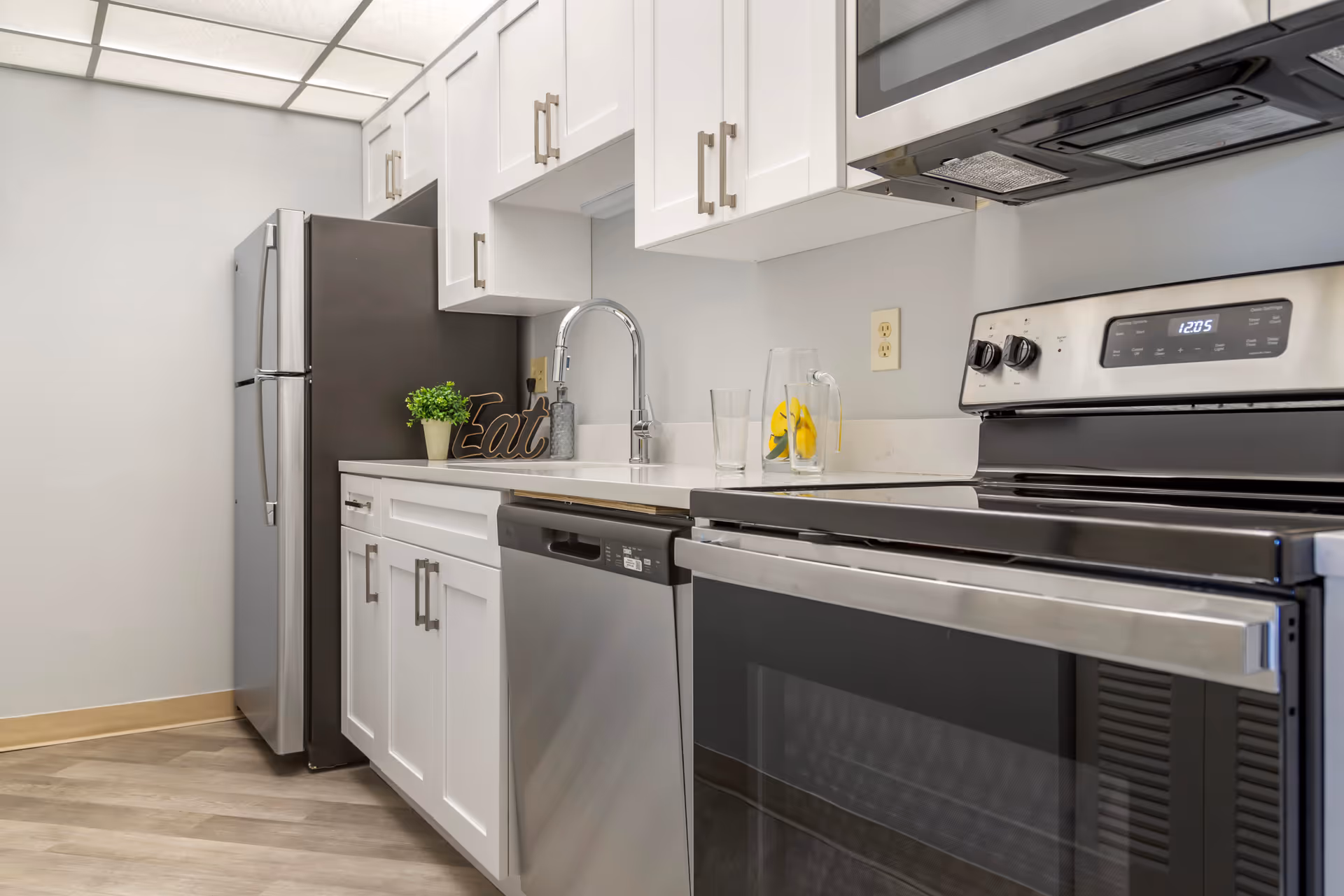 Modern kitchen with stainless steel refrigerator, oven, dishwasher, white cabinets and a sink on a light countertop.
