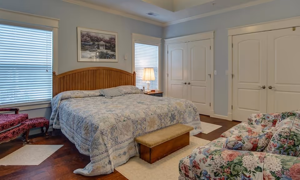 Bright bedroom with a large bed covered in a patterned quilt, a floral armchair, nightstand with a lamp, and double closet doors.