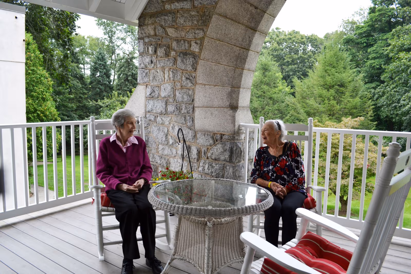 Two elderly women sitting and chatting on white rocking chairs around a glass-top wicker table on a covered porch with a stone arch and green trees in the background.