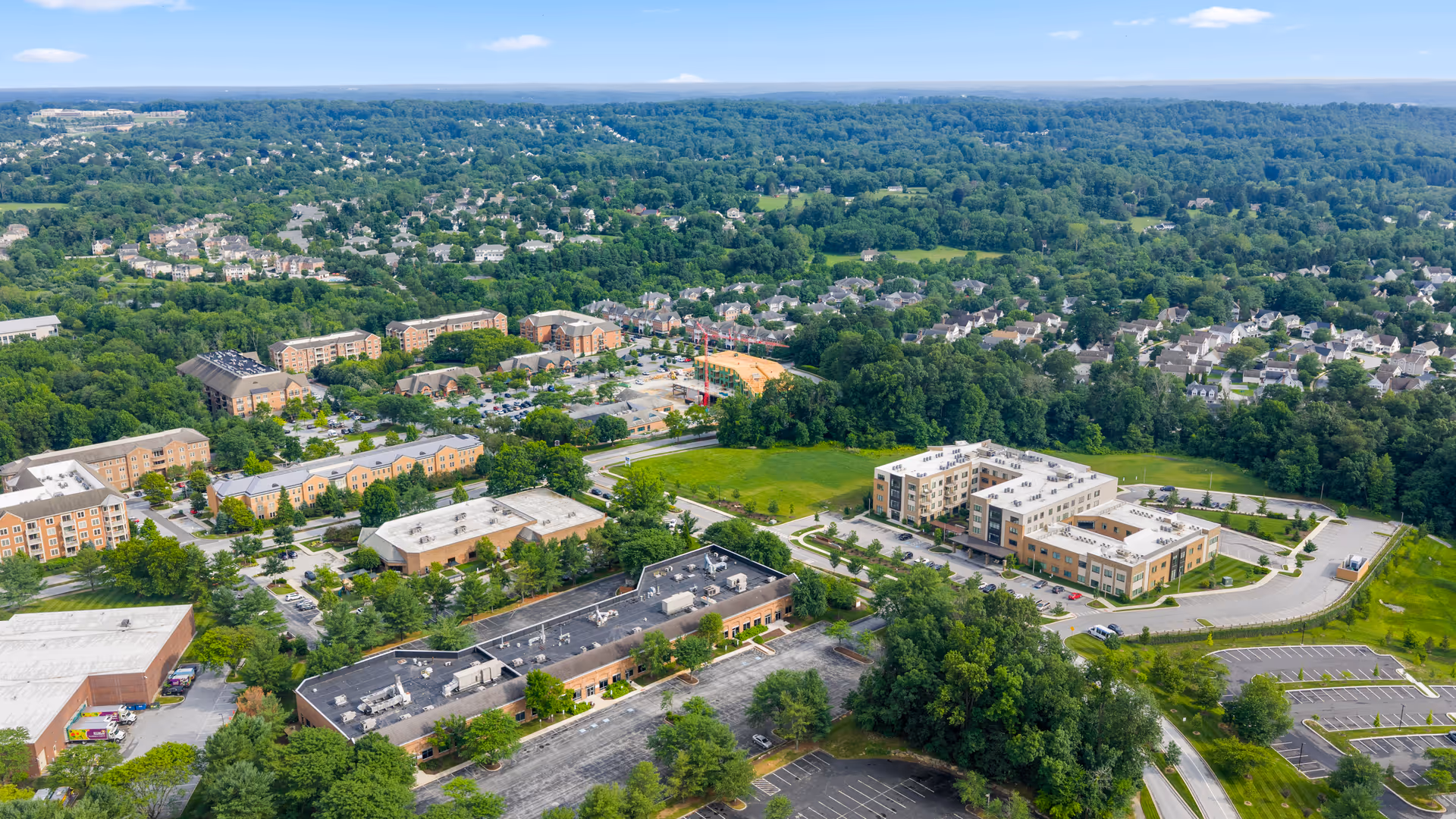 Aerial view of a suburban area featuring multiple residential buildings, parking lots, and green spaces surrounded by dense trees and a distant horizon under a partly cloudy sky.