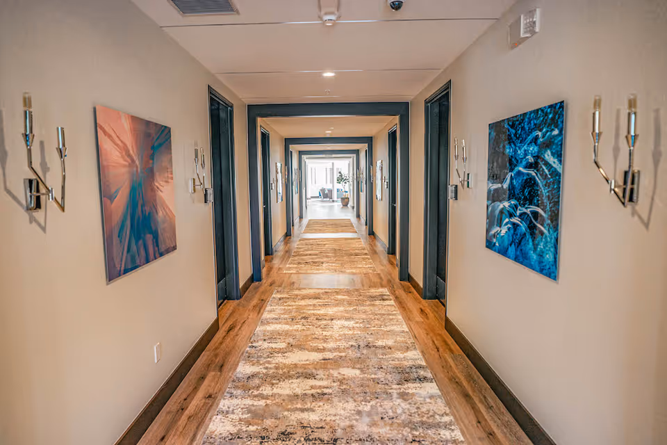 A long, well-lit hallway in a senior living facility with wooden floors and a patterned runner rug. The walls are decorated with modern artwork and wall-mounted candle holders. Multiple doorways line both sides of the hallway, and natural light is visible at the far end.
