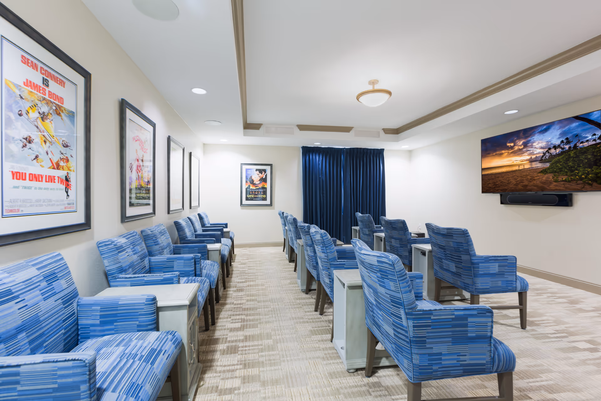 A small theater or media room with two rows of blue patterned armchairs facing a wall-mounted flat screen TV displaying a beach sunset scene. The room has beige walls adorned with framed vintage movie posters, recessed ceiling lights, and a ceiling light fixture. There are small side tables between the chairs and dark blue curtains covering a window at the back.