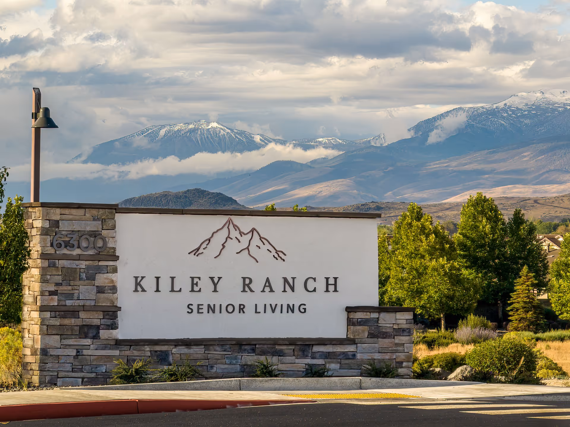 Stone and stucco sign for Kiley Ranch Senior Living with mountain logo, set against a backdrop of green trees and snow-capped mountains under a cloudy sky.