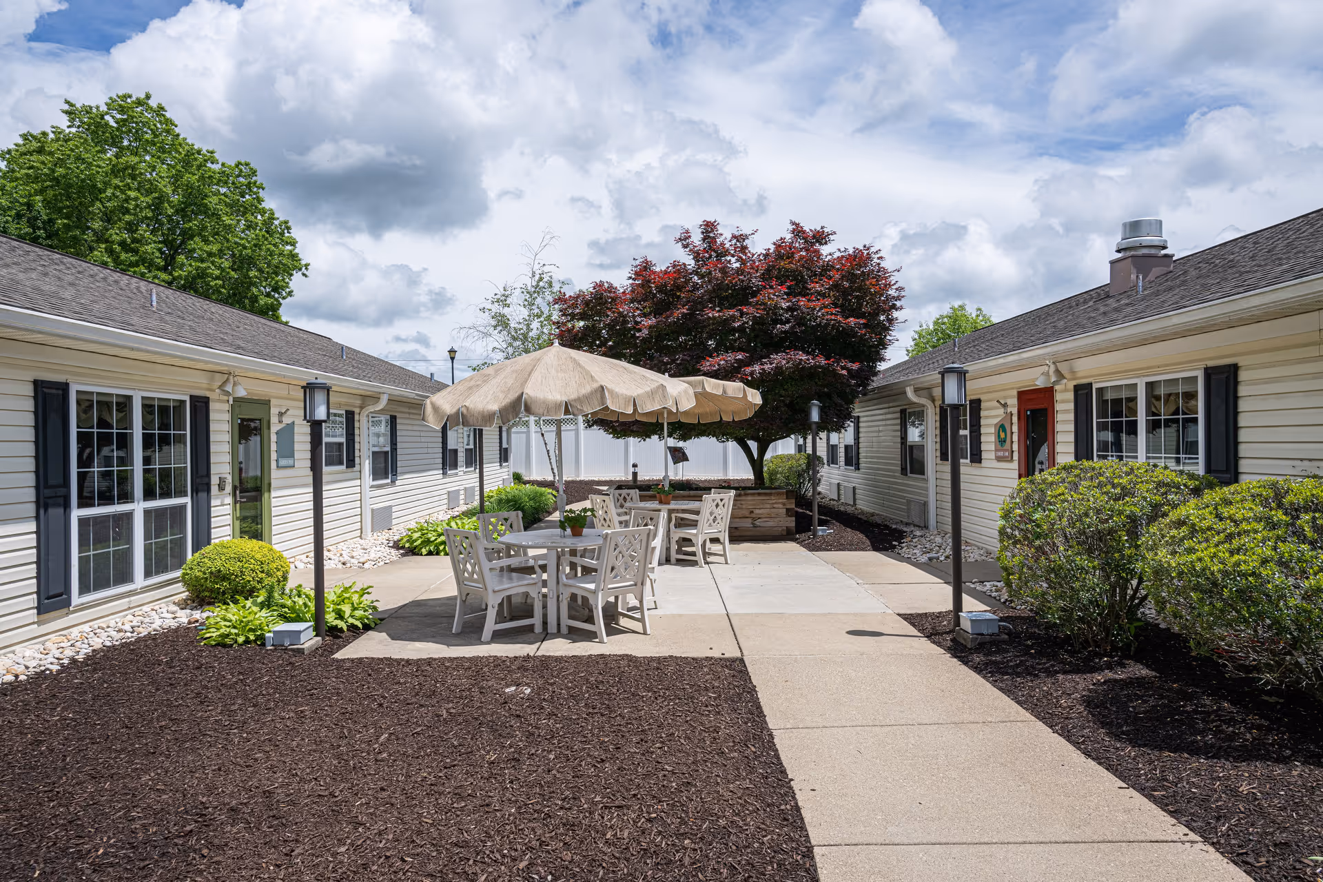 Outdoor courtyard area between two single-story buildings with beige siding and black shutters. The courtyard features a concrete walkway, patio tables with umbrellas, chairs, landscaped bushes, and a tree with red leaves under a partly cloudy sky.