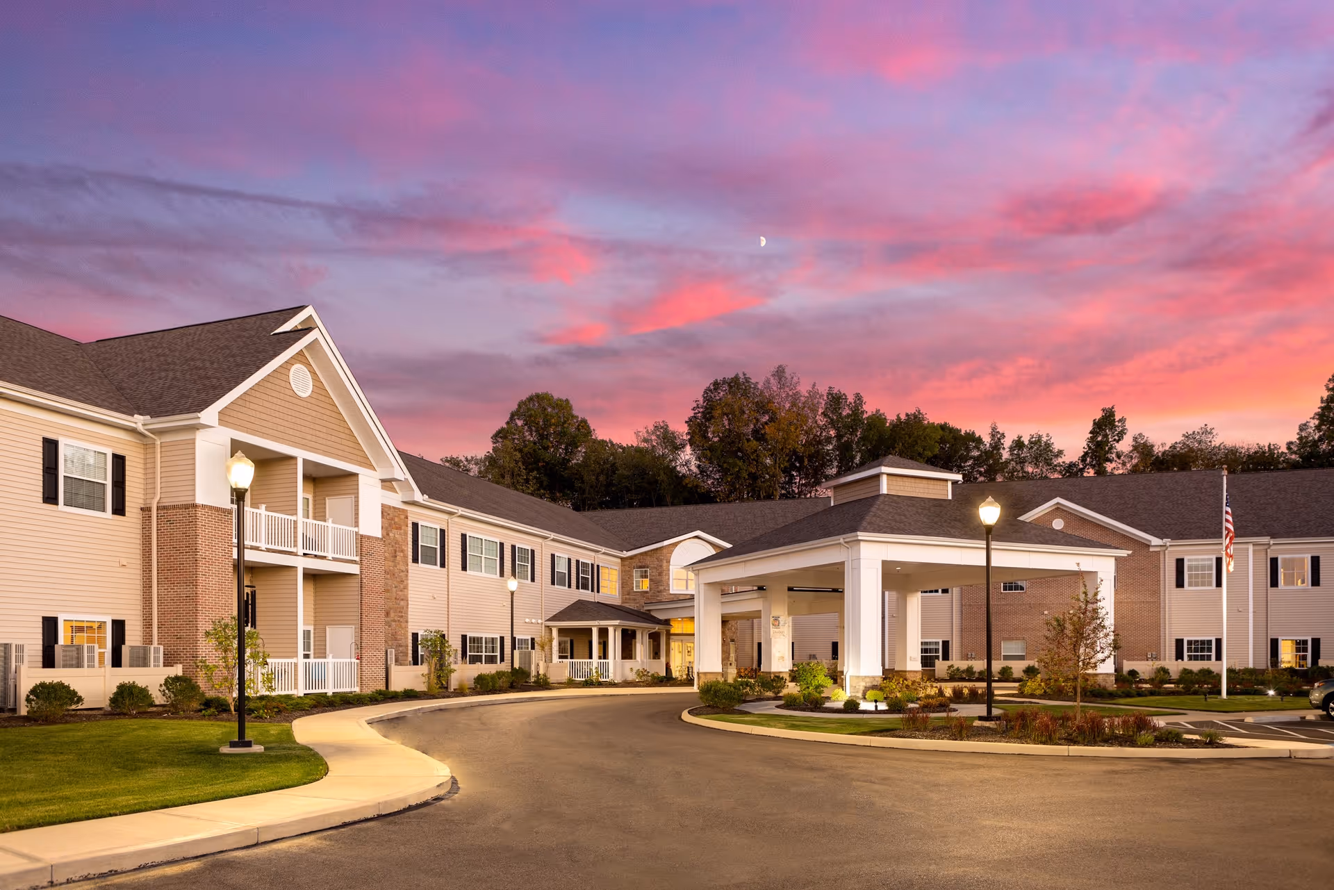 Senior living facility exterior with a circular driveway, lit entrance, and pink sunset sky.