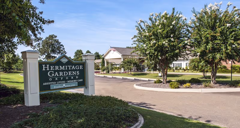 Exterior view of Hermitage Gardens Oxford assisted living and memory care facility with a green sign in the foreground and a driveway leading to the building entrance surrounded by trees and landscaping under a clear blue sky.