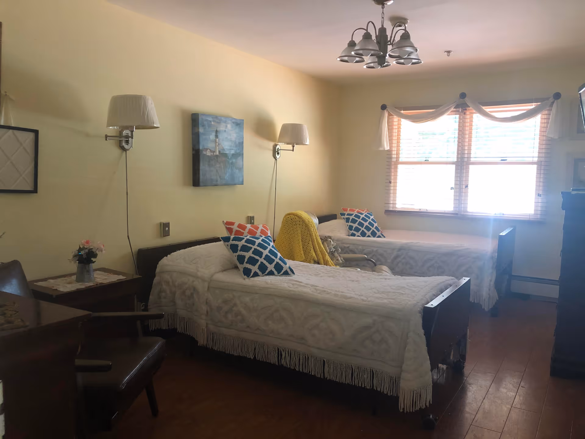 A bedroom with two single beds covered in white patterned bedspreads, each with a colorful pillow. There is a yellow blanket draped over a chair between the beds. The room has wooden flooring, a window with blinds and a decorative curtain, wall-mounted lamps above each bed, a painting on the wall, and a wooden dresser with a TV on top.