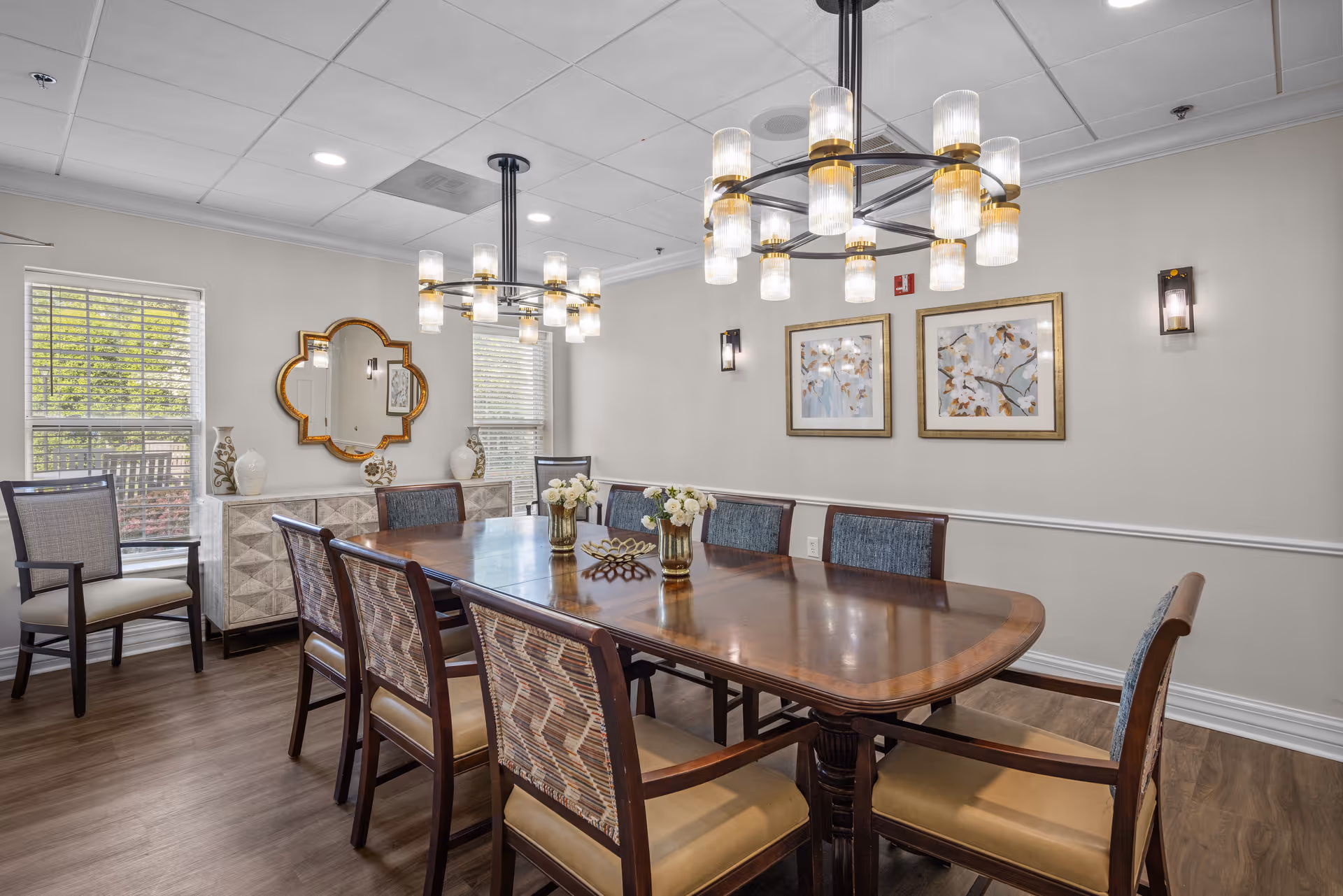 A well-lit dining room with a large wooden dining table surrounded by eight chairs. The room features two modern chandeliers with multiple glass shades, two windows with blinds, a decorative mirror on the wall, two framed floral artworks, and a sideboard with vases and decorative items. The floor is wooden, and the walls are painted light beige with white trim.
