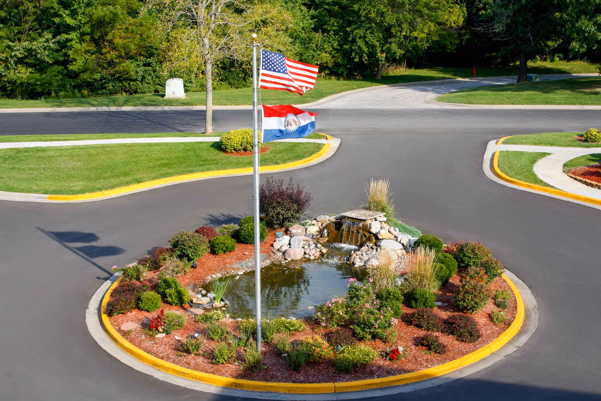 A landscaped roundabout with a small pond and waterfall feature in the center, surrounded by various shrubs and flowers. Two flags, the American flag and a state flag, are flying on a flagpole in the middle of the roundabout. The area is bordered by a paved road and green grass with trees in the background.