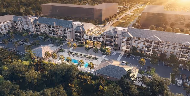 Aerial view of a senior living facility with multiple connected buildings surrounding a central outdoor pool area with lounge chairs and palm trees, parking lots on either side, and lush greenery in the foreground during sunset.