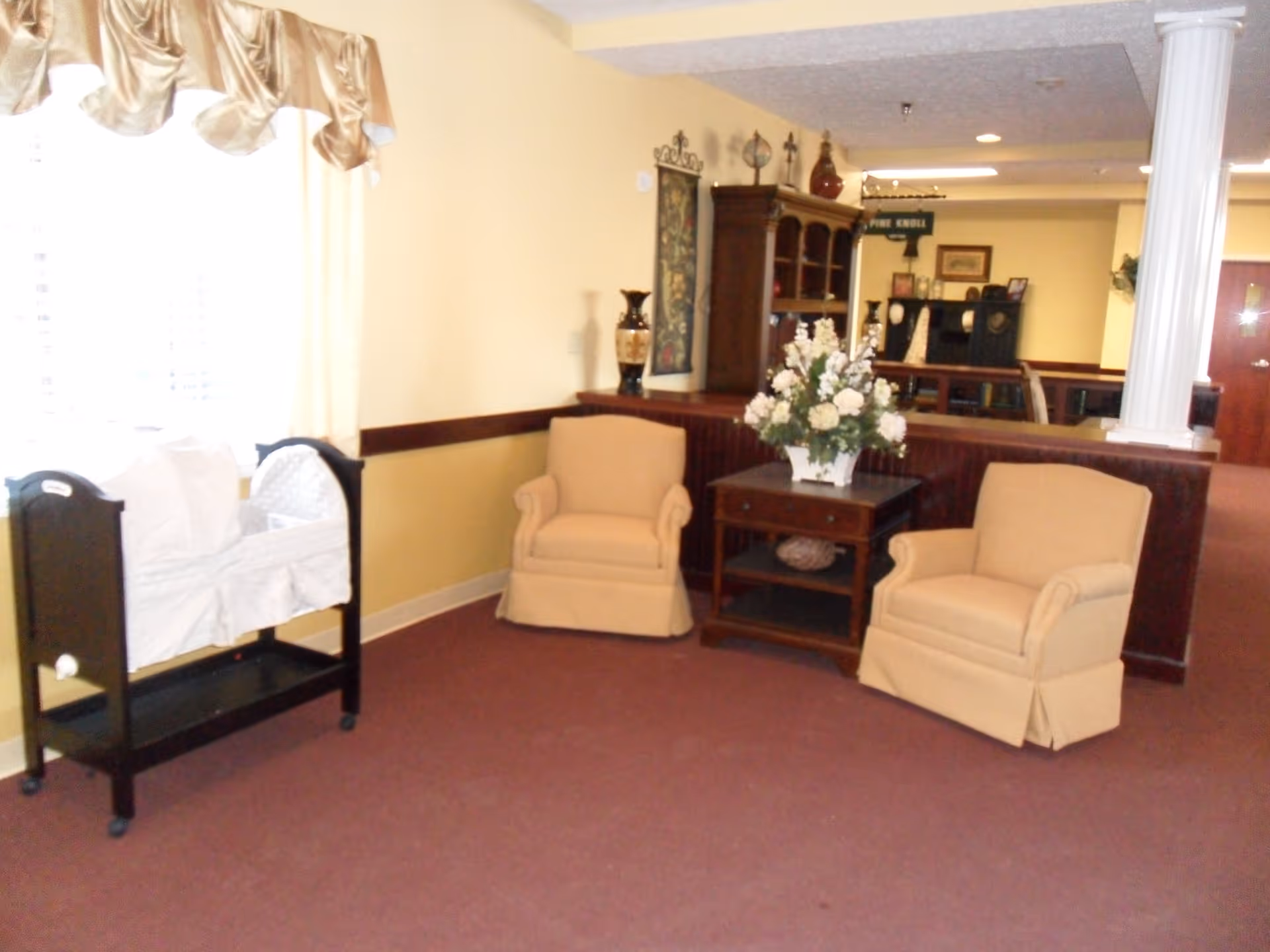 Carpeted seating area with two beige armchairs and a side table topped with a floral arrangement beside a window and small cradle stand.