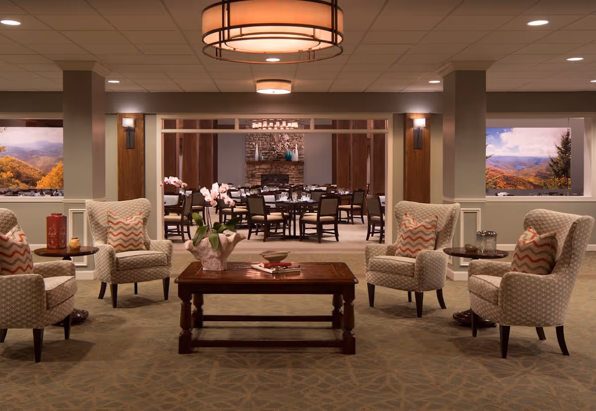 A cozy sitting area with four patterned armchairs arranged around a wooden coffee table with a flower vase and books. Behind the sitting area is a dining room with multiple tables and chairs, a stone fireplace, and decorative vases. Large windows on either side show scenic views of autumn-colored mountains.