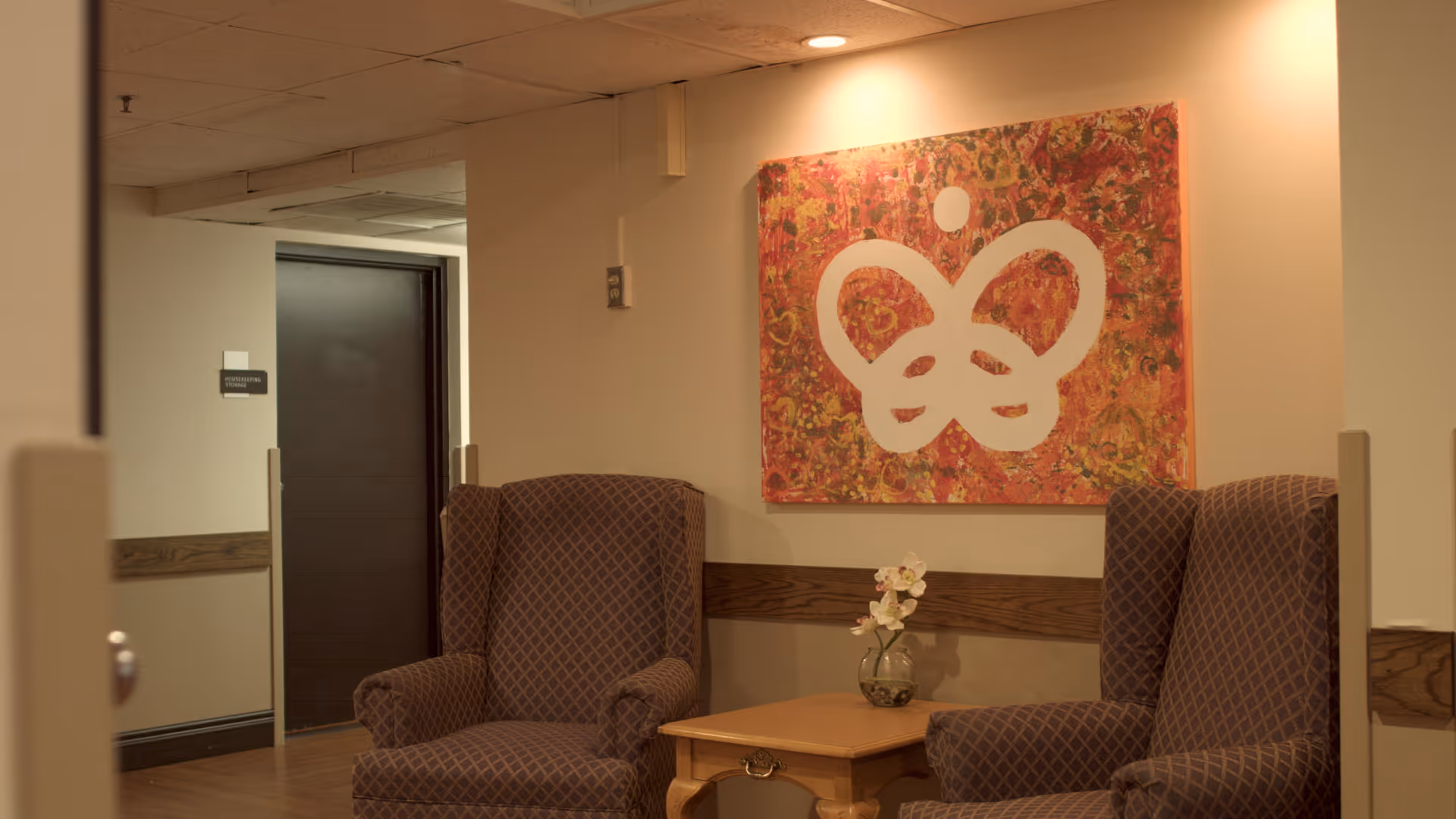 A cozy seating area in a hallway with two patterned armchairs facing each other and a small wooden table with a glass vase holding white flowers between them. A colorful abstract painting with a white butterfly-like design hangs on the wall behind the chairs.