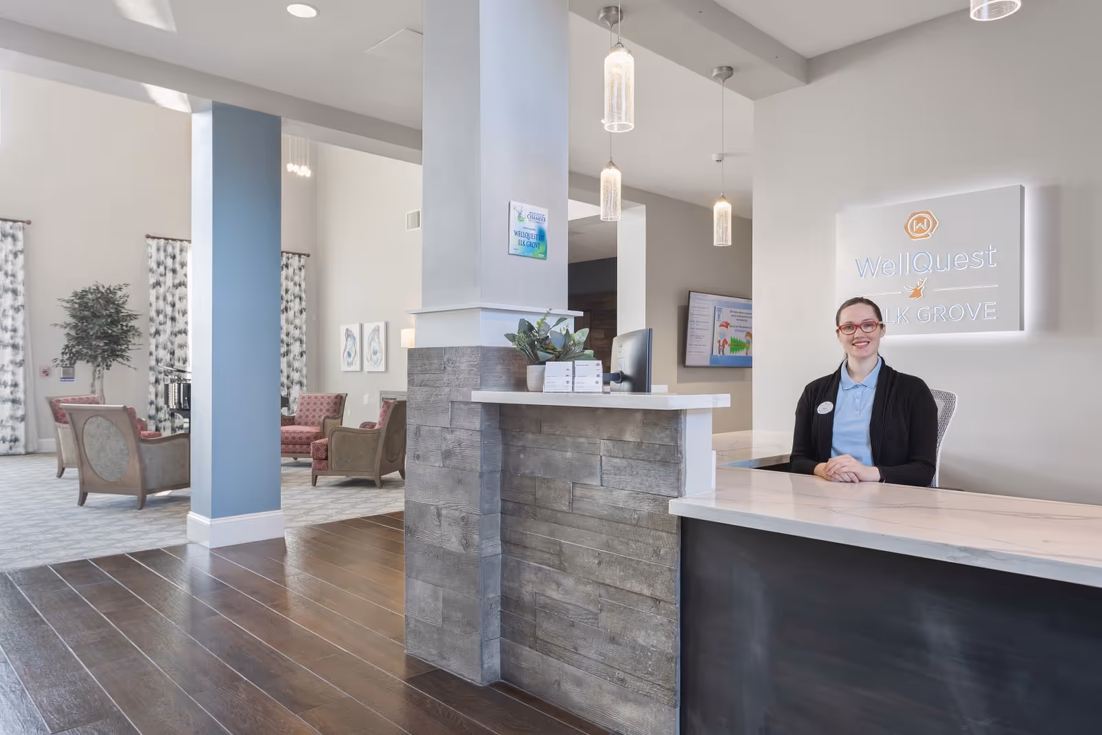 Reception area of WellQuest of Elk Grove featuring a smiling receptionist sitting behind a modern desk with a wood and marble finish. The background includes a seating area with chairs, tall windows with patterned curtains, and a sign on the wall displaying the WellQuest of Elk Grove logo.