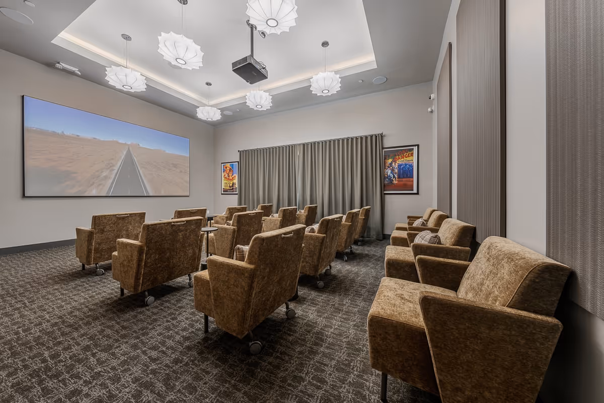 A small theater room with two rows of brown upholstered chairs on wheels facing a large screen displaying a road through a desert landscape. The room has a patterned carpet, modern ceiling lights, and framed movie posters on the walls with gray curtains covering the windows.