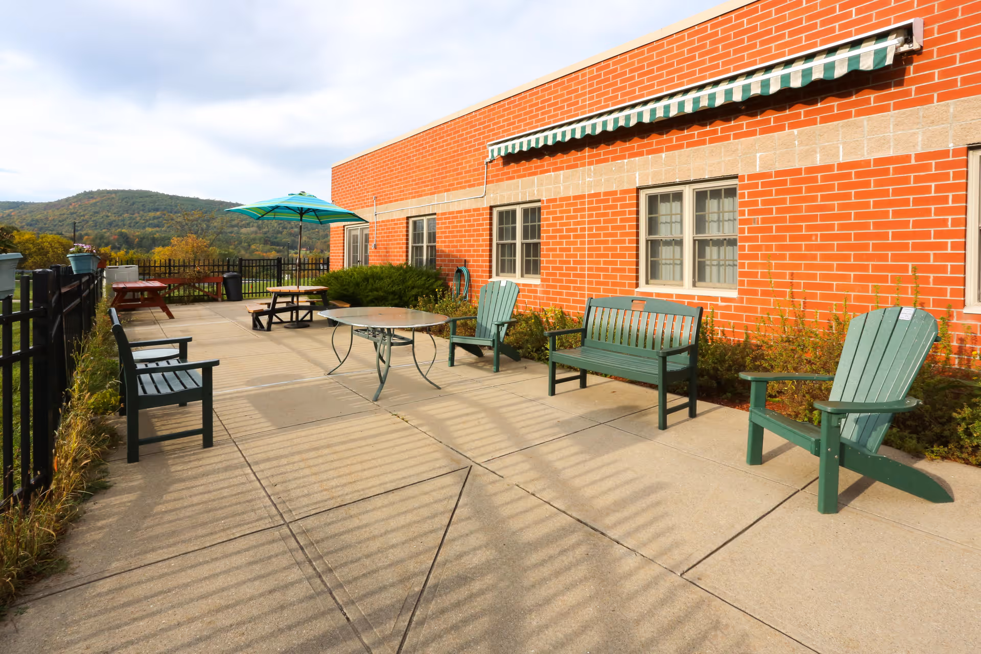 Outdoor patio area with green Adirondack chairs, benches, a round metal table, and picnic tables with umbrellas next to a red brick building. There is a black metal fence along the patio and a view of tree-covered hills in the background under a partly cloudy sky.