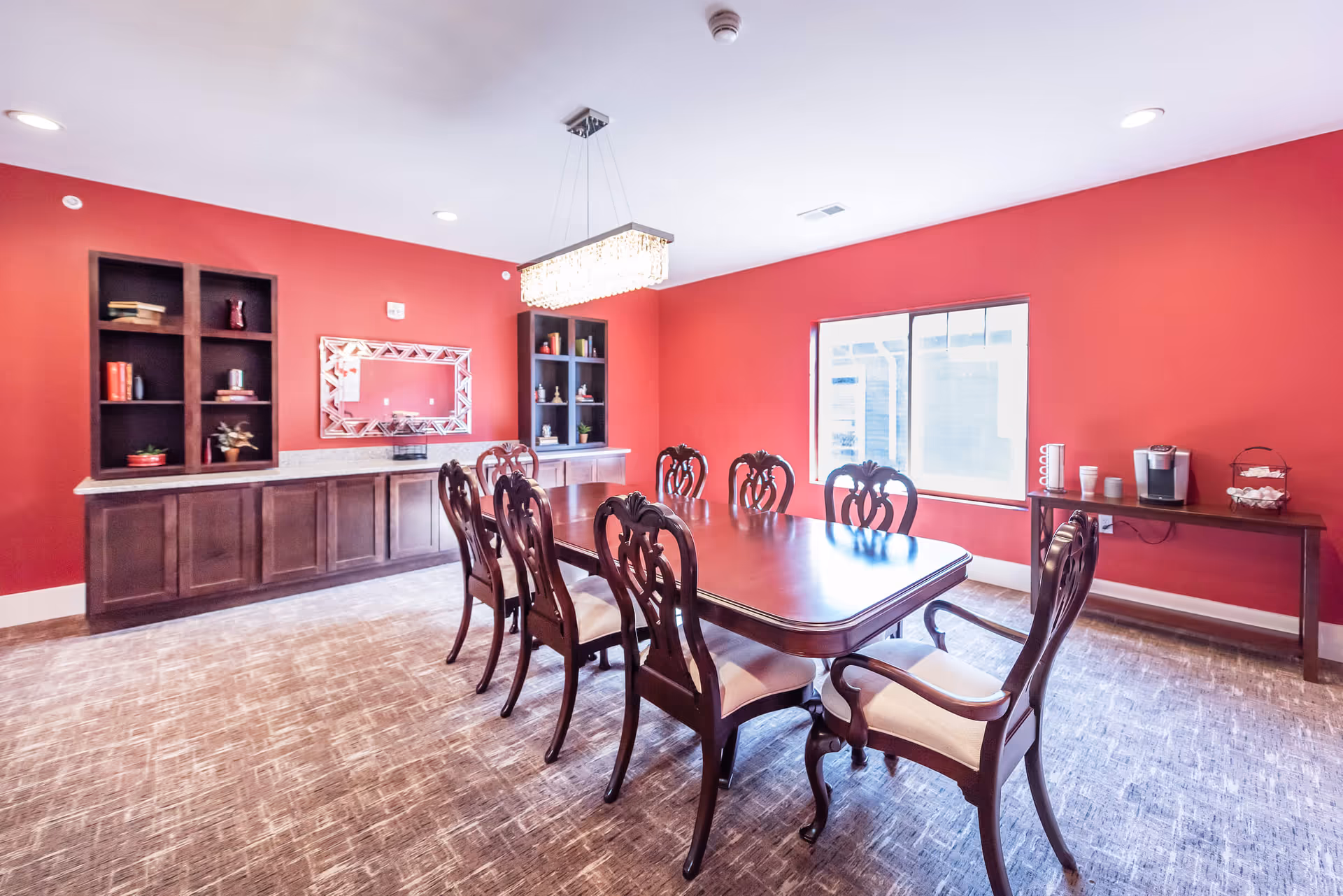 A dining room with red walls and a large wooden dining table surrounded by eight ornate wooden chairs with light cushions. There is a modern rectangular chandelier hanging above the table. Against one wall, there are built-in dark wooden shelves and cabinets with decorative items and books. A large window lets in natural light, and a side table holds a coffee maker and cups.