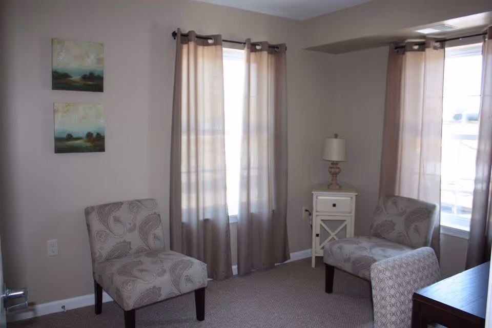 A small sitting area with two patterned armchairs facing each other near two windows with beige curtains. Between the chairs is a small white side table with a lamp on it. Two landscape paintings hang on the wall to the left.