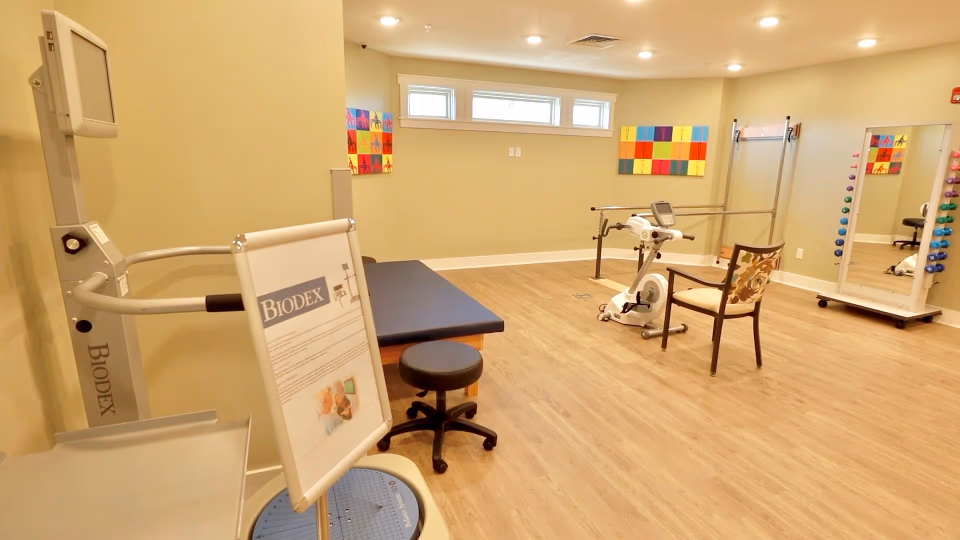 Small therapy/fitness room with exercise equipment, a treatment table, chair, and colorful wall art.