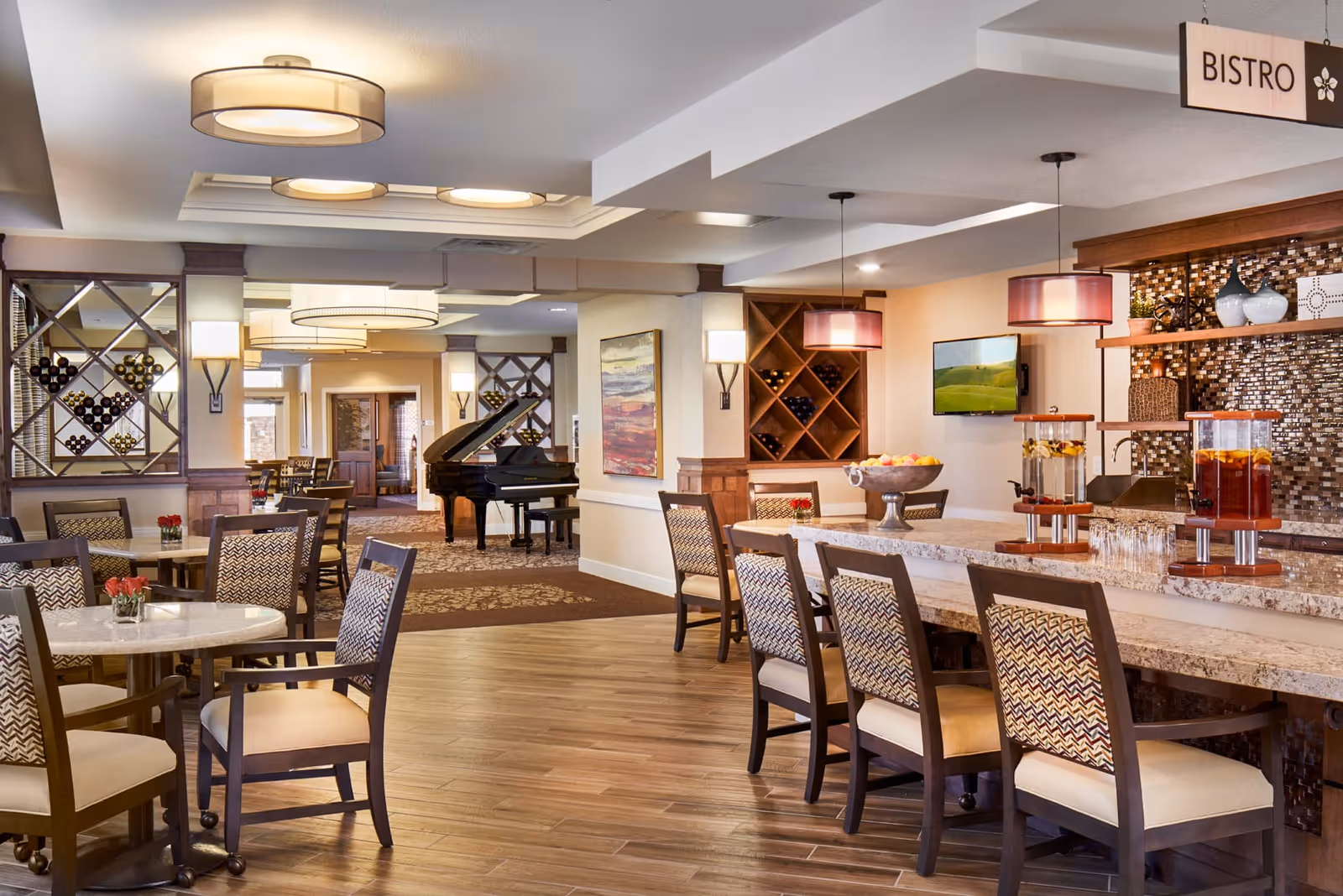 Interior view of a bright and elegant bistro area in an assisted living facility featuring a long marble counter with chairs, beverage dispensers, small round tables with chairs, a grand piano in the background, wine racks on the walls, and modern ceiling lights.