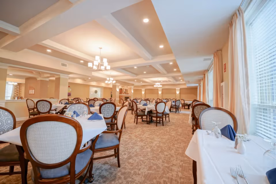 Spacious dining room with multiple tables covered in white tablecloths, set with blue napkins, glassware, and silverware. The room features elegant wooden chairs with patterned upholstery, large windows with curtains allowing natural light, and ceiling lights with chandeliers.