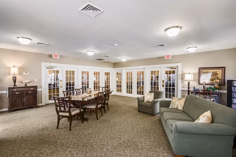 A spacious senior living common area with beige walls and carpeted floor. The room features a long wooden dining table with eight chairs, a green sofa and armchair with floral cushions, a wooden sideboard with a lamp and decorative items, and a small desk with a lamp and framed picture. Multiple white-framed glass doors line the walls, and ceiling lights illuminate the room.
