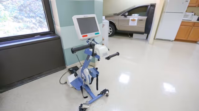 A medical exercise machine with a digital screen is positioned near a window inside a room. In the background, there is a partial view of a vehicle with an open door and a kitchen area with a refrigerator and wooden cabinets.