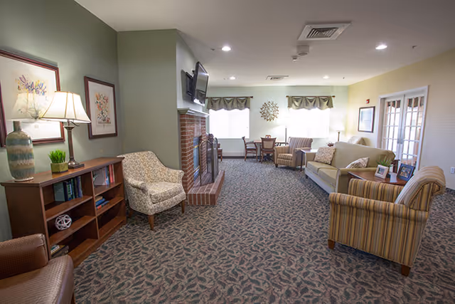A cozy living room area in a senior living facility featuring a patterned carpet, a brick fireplace with a mounted TV above it, several upholstered chairs and a sofa, a wooden bookshelf with books and decorative items, framed artwork on the walls, and large windows letting in natural light.