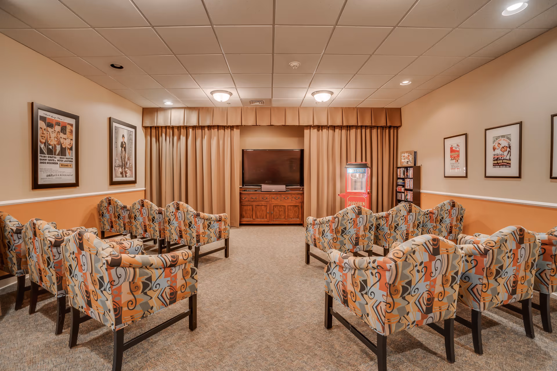 A cozy movie room with patterned armchairs arranged in rows facing a flat-screen TV on a wooden cabinet. There is a popcorn machine and a shelf with DVDs on the right side. The walls are decorated with framed movie posters, and beige curtains cover the back wall.