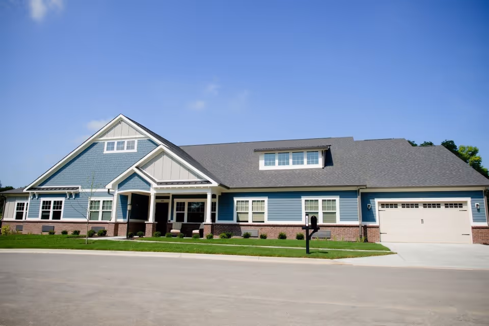 Exterior view of a single-story residential building with blue siding, white trim, and a gray shingled roof under a clear blue sky. The building has multiple windows, a covered front porch with seating, a garage on the right side, and a small lawn with a mailbox in front.