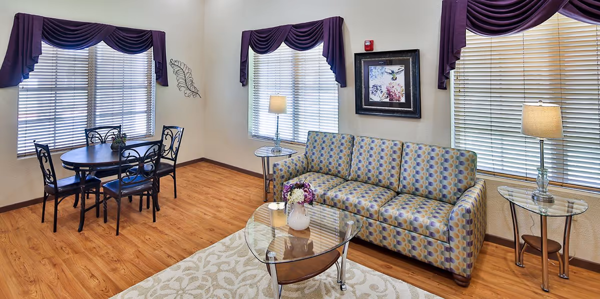 A cozy living room area with a patterned sofa, glass coffee table with a flower vase, two side tables with lamps, and a round dining table with four chairs. The room has wooden flooring, three windows with blinds and purple valances, and a framed floral artwork on the wall.