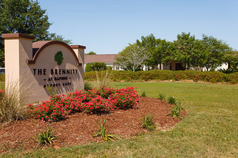Outdoor view of The Brennity at Daphne Assisted Living & Memory Care sign surrounded by landscaped flowers and greenery with the facility building and trees in the background under a clear blue sky.