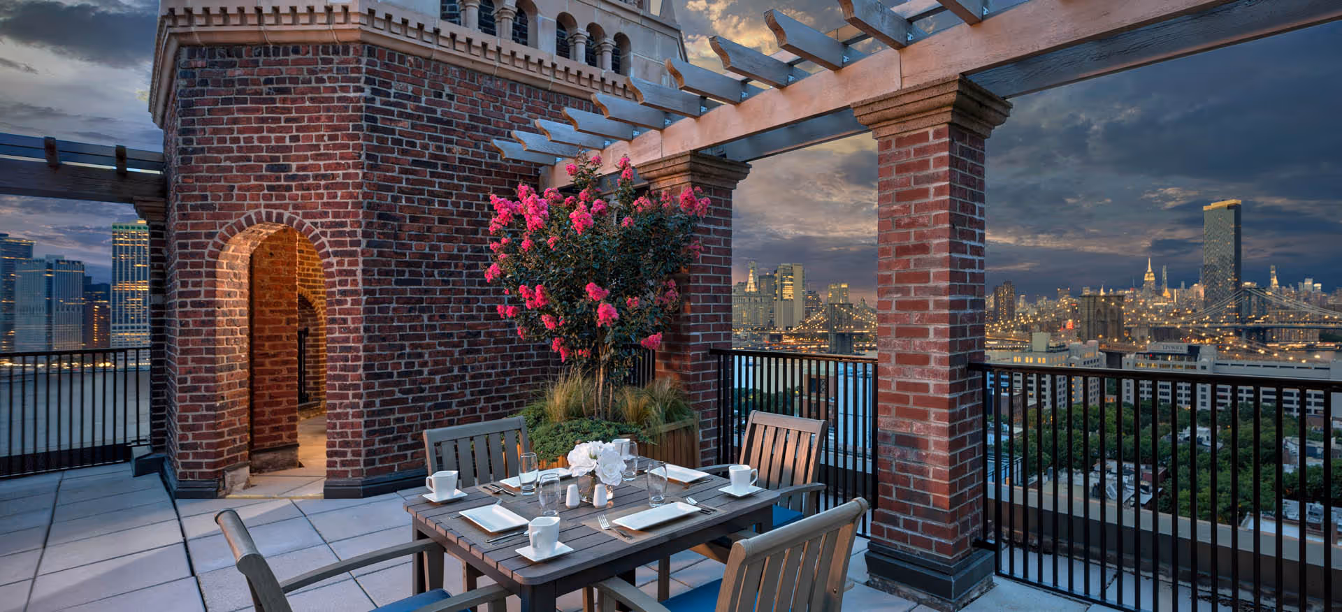 Outdoor terrace with a dining table set for four, surrounded by wooden chairs. The terrace features brick columns and a pergola overhead. A flowering plant with bright pink blossoms is placed near the brick wall. The background shows a city skyline at dusk with illuminated buildings and bridges.