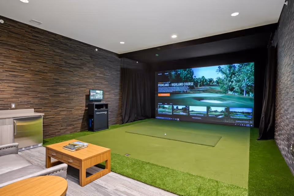 Indoor golf simulator room with a large screen displaying a golf course, artificial green turf flooring, a small wooden coffee table with books, a leather chair, and a mini fridge against a textured dark wall.