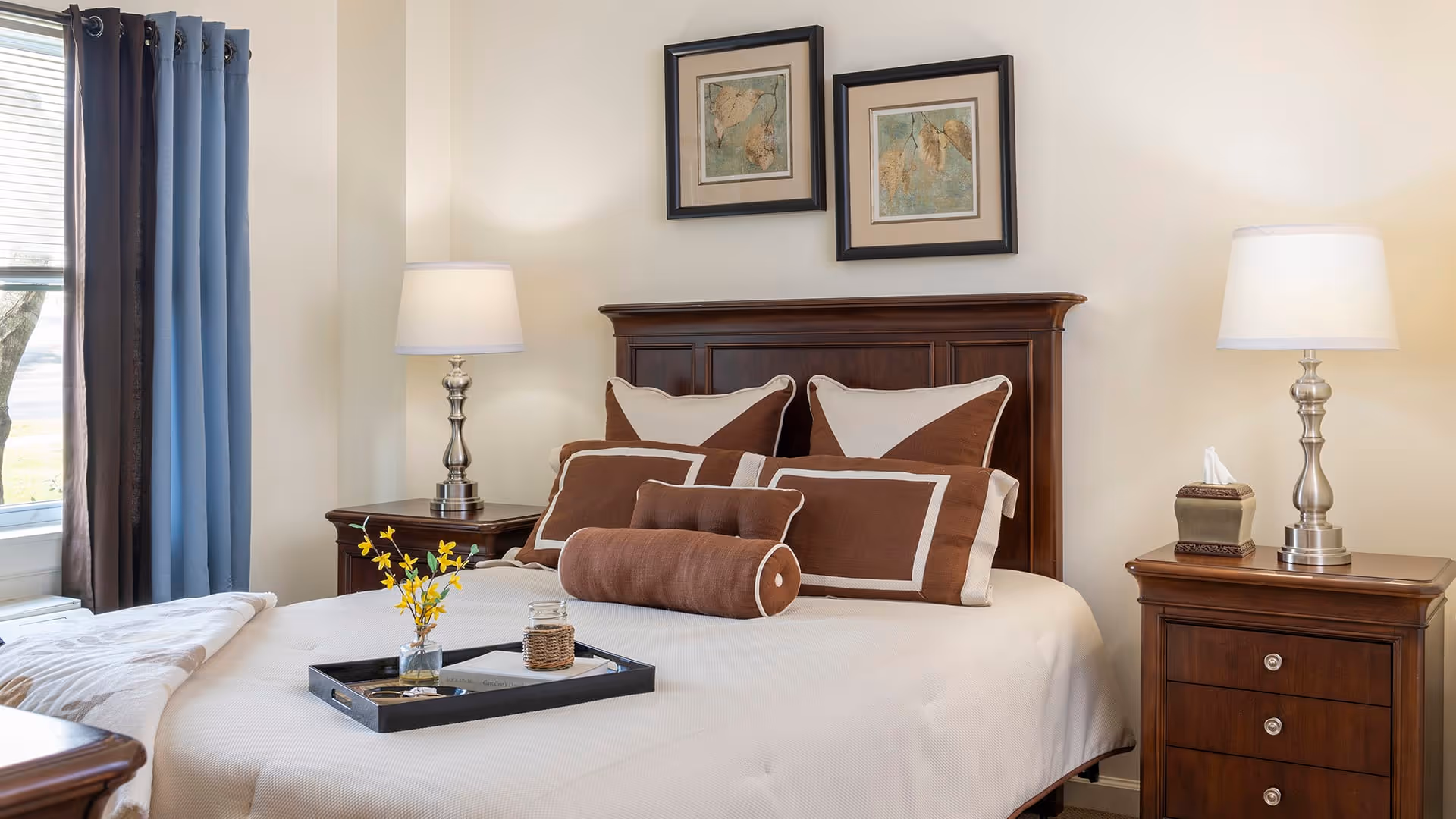 A neatly made bed with brown and beige pillows and a matching bolster pillow in a bedroom. The bed has a dark wooden headboard and is flanked by two wooden nightstands, each with a silver lamp. There are two framed maps hanging on the wall above the bed. A window with blue and brown curtains is visible on the left side, and a tray with a small vase of yellow flowers and a candle is placed on the bed.