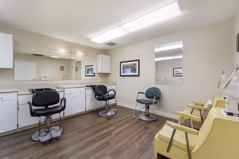 Small salon-style room with barber chairs, mirrors, cabinets and yellow waiting chairs on wood flooring.