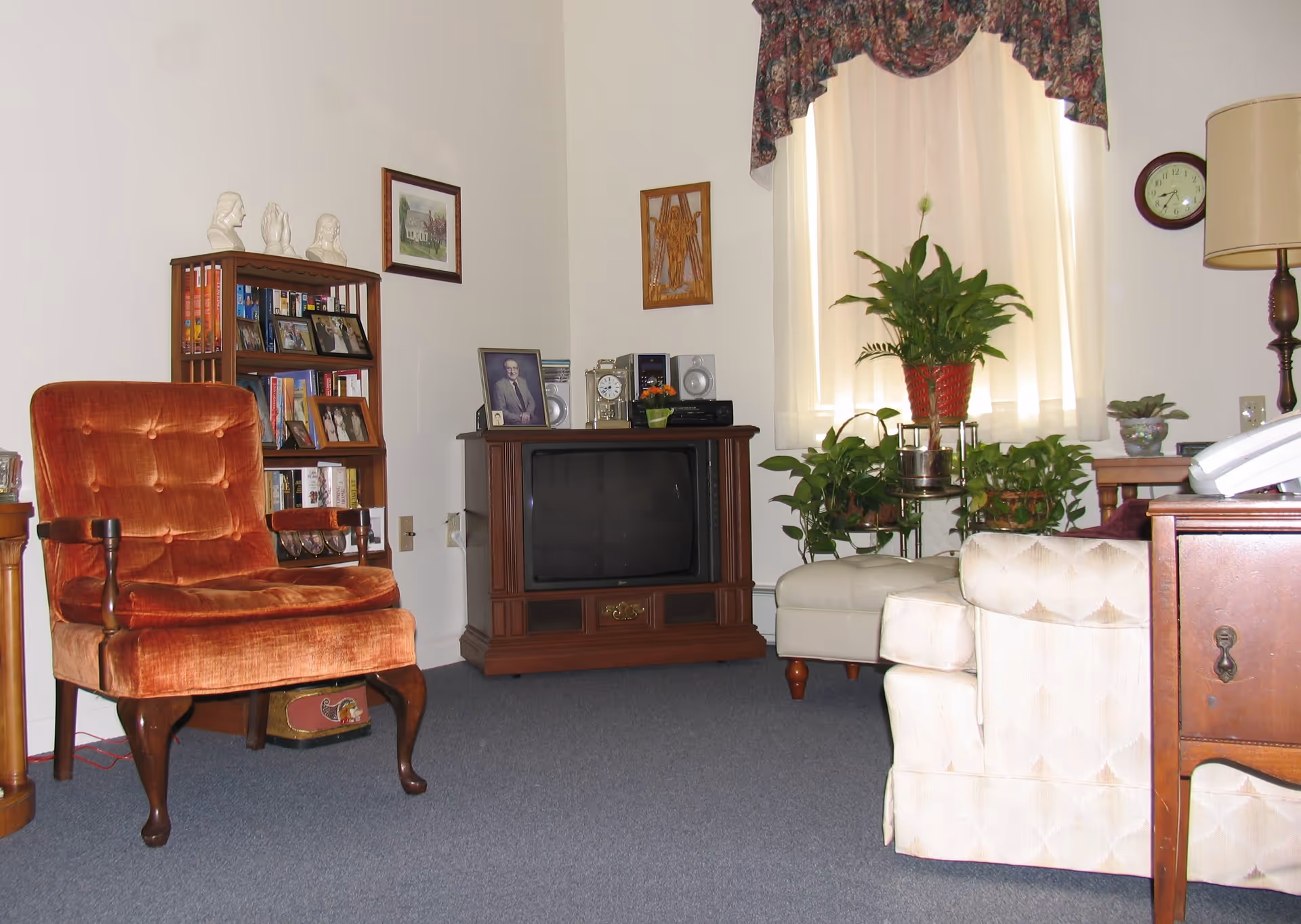 A cozy living room with a vintage orange upholstered armchair, a wooden bookshelf filled with books and framed photos, a wooden TV stand with an old-style television, a clock, stereo speakers, and decorative items. There is a beige armchair partially visible, a wooden side table with a lamp, several potted plants near a window with cream curtains and a floral valance, and framed artwork on the walls.
