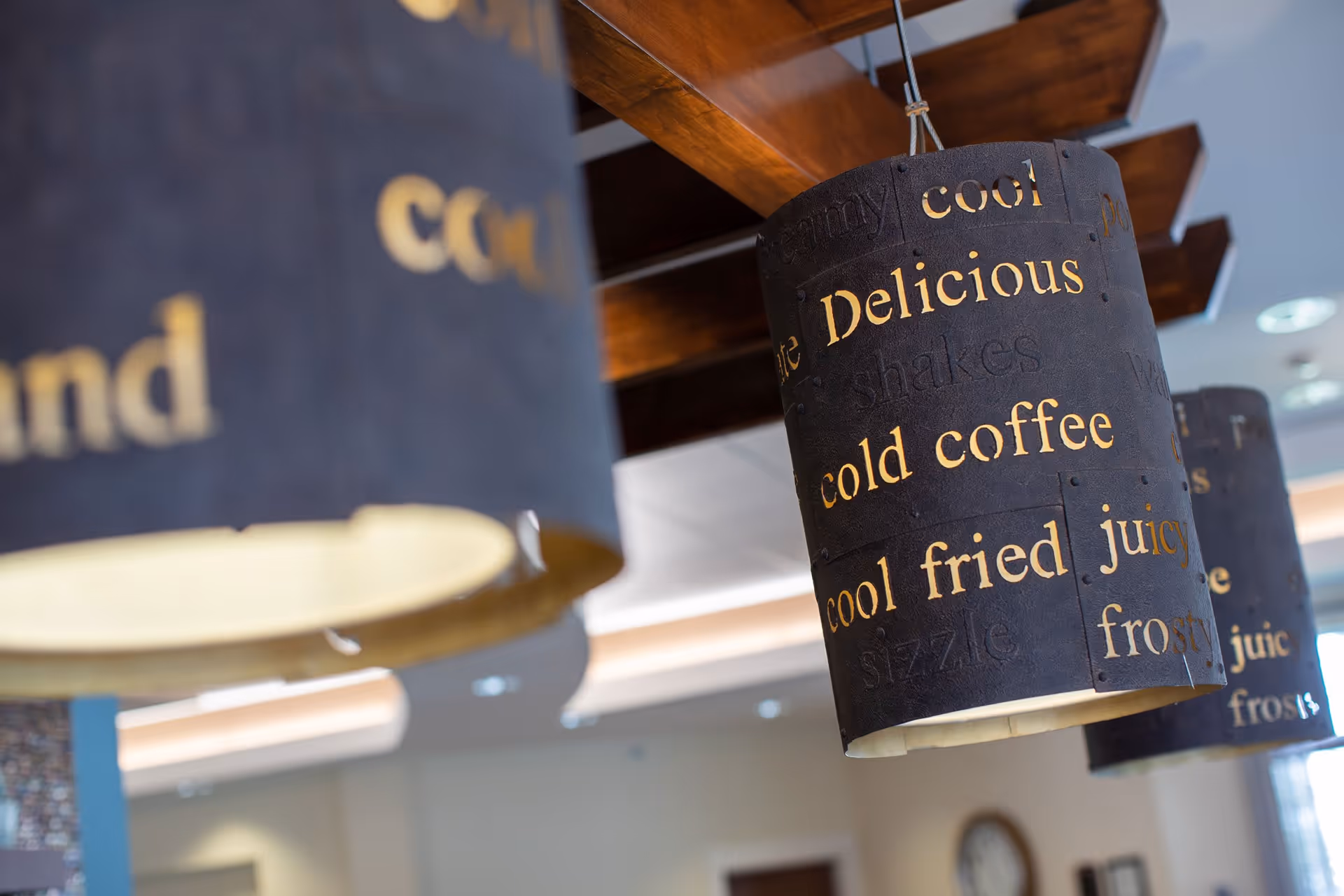 Close-up of decorative hanging light fixtures with cut-out words such as 'Delicious', 'cold coffee', 'cool fried', and 'juicy' illuminated from inside, in an indoor setting with wooden beams on the ceiling.