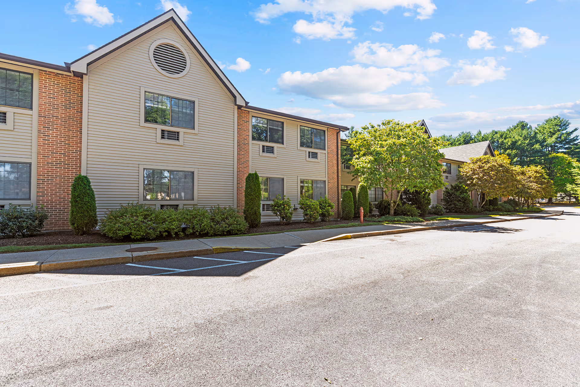 Exterior view of a two-story senior living facility building with beige siding and brick accents, surrounded by neatly trimmed bushes and trees under a blue sky with scattered clouds.